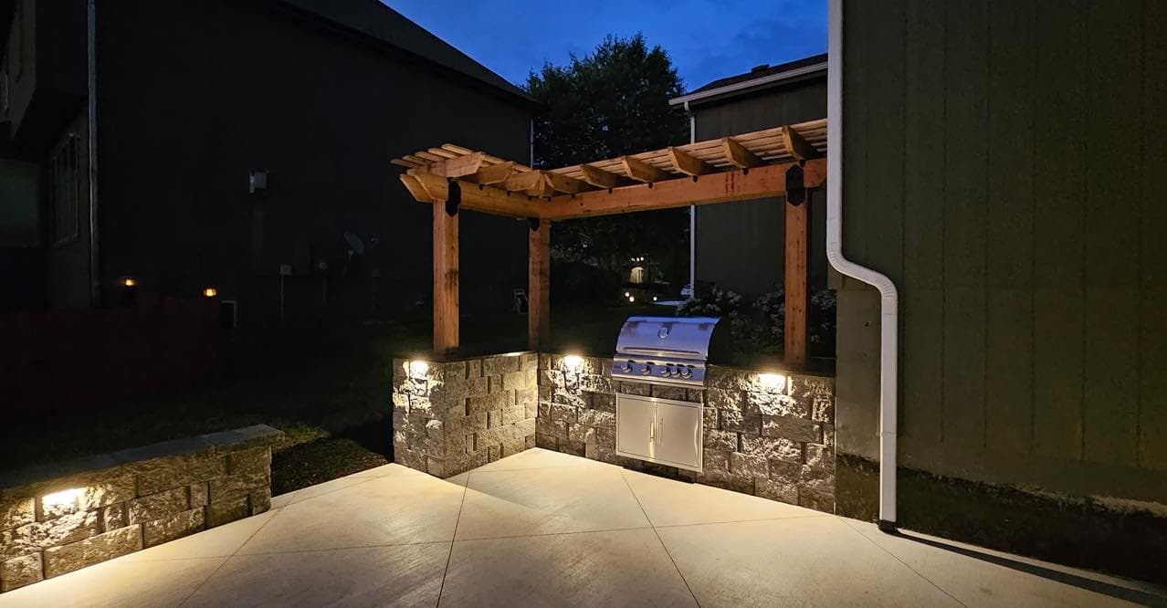 Modern outdoor kitchen with a stainless steel grill under a wooden pergola at night.